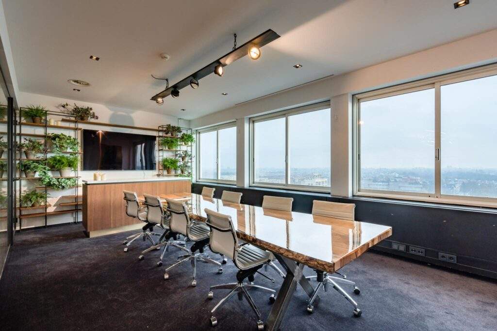 Vergaderruimte aan het Koningin Wilhelminaplein met een grote houten tafel, moderne stoelen, veel daglicht en een groen aangeklede wand met televisie.